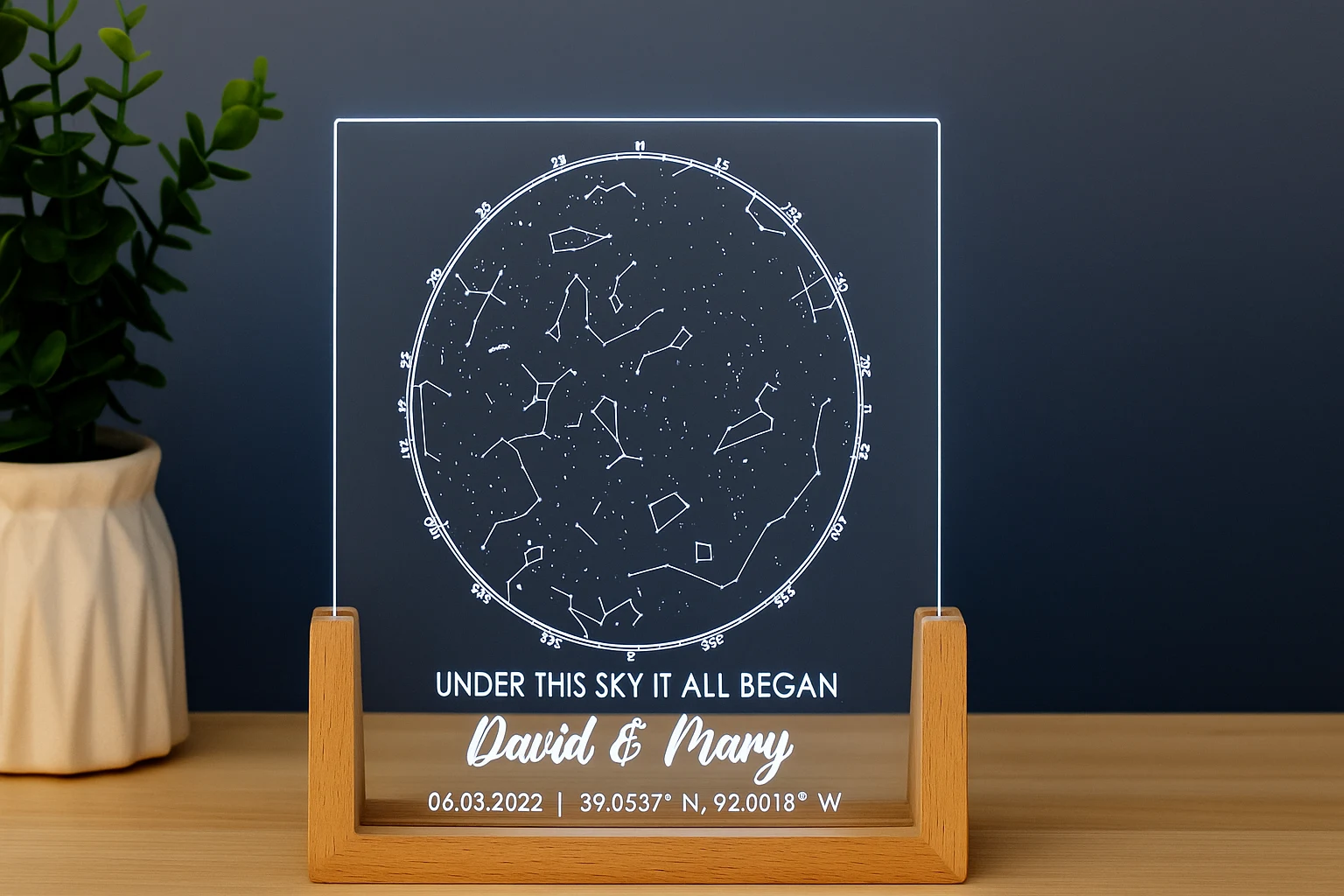 LED-lit acrylic star map with wooden base and personalized names displayed on a wooden table beside a white ceramic plant pot.