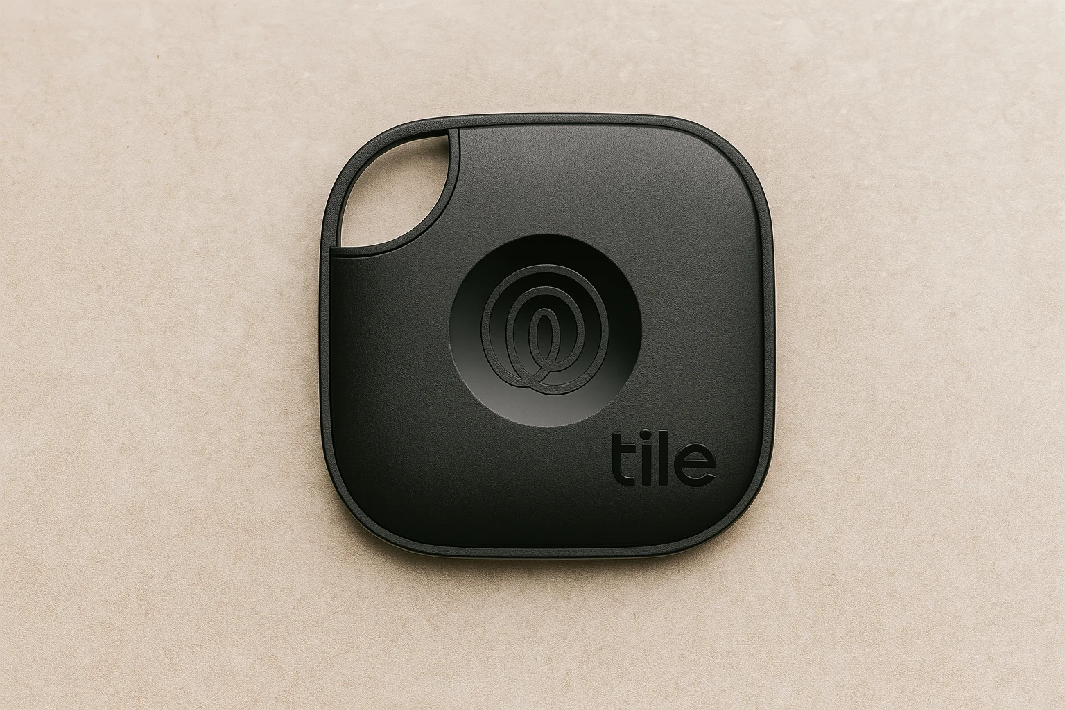 Close-up of a black Tile Bluetooth tracker lying on a beige surface, designed to help users locate lost keys, bags, or devices.
