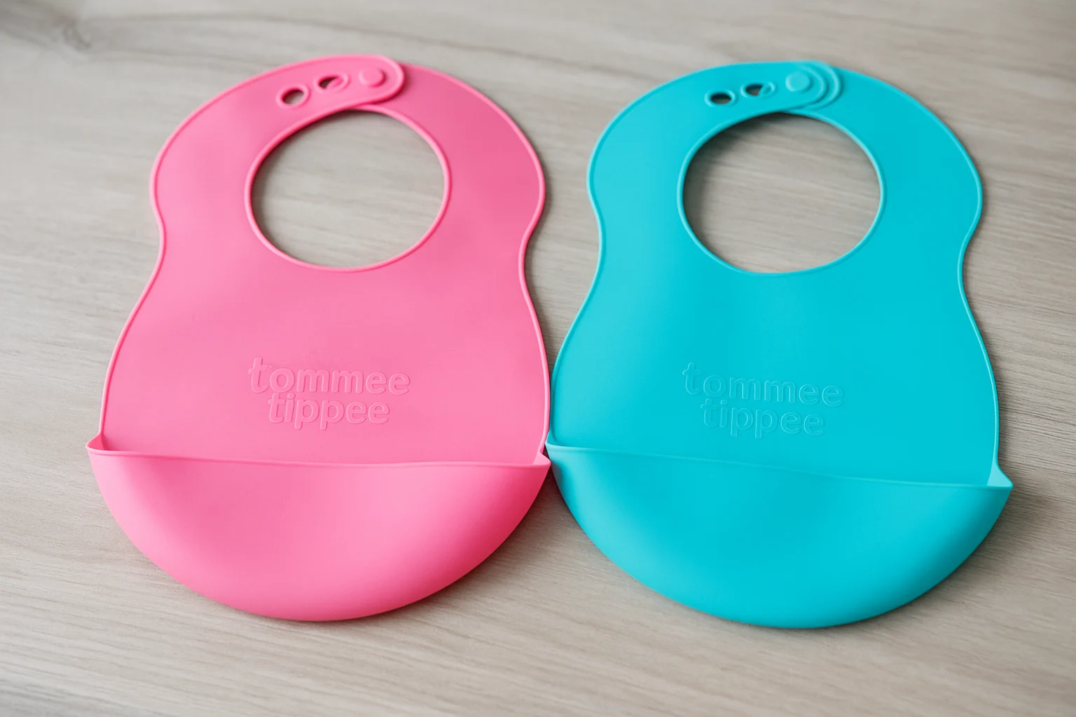 Two Tommee Tippee silicone bibs in pink and turquoise on a wooden surface